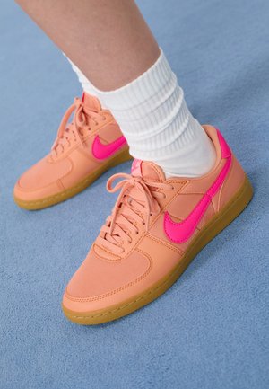 Fødder iført ferskenfarvede sneakers med lyserøde Nike-swoosh-logoer og hvide ribstrikkede crew-sokker på en lyseblå overflade.
