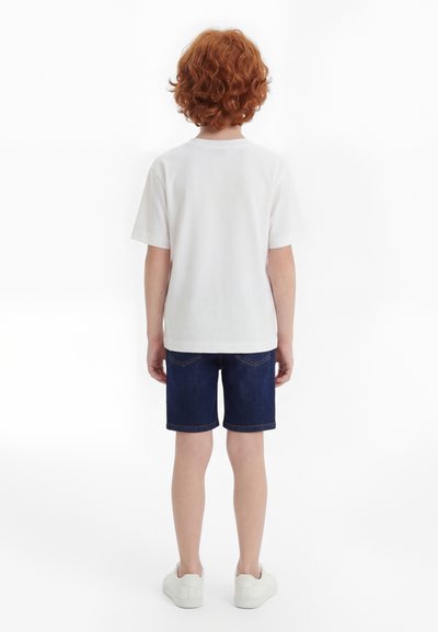 Laps lokkis punaste juustega, kes kannab lihtsat valget T-särki, tumesiniseid teksapükse ja valgeid tenniseid, seisab valgel taustal selg vaataja poole.