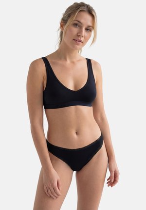 Frau trägt einen schwarzen Soft-Cup-BH und passende schwarze Low-Rise-Slips, steht vor einem schlichten weißen Hintergrund.