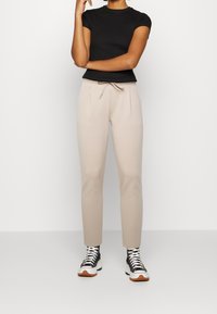 Béžové joggingové kalhoty s elastickým pasem, bočními kapsami a elastickými manžetami, zkombinované s upnutým černým tričkem a vysokými teniskami.