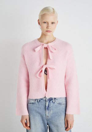 Lyserød ribbet cardigan med to forreste sløjfer og åben midte. Lange ærmer og blød tekstur, parret med lyseblå denim jeans.