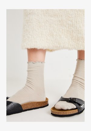 Zwarte slide sandalen met een textuurafwerking, een kurken voetbed en een band met een gesp, gedragen met beige geribbelde sokken.