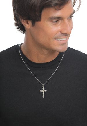 Mann trägt schwarzes Hemd und silberne Kreuzkette, lächelt und schaut seitlich vor weißem Hintergrund.