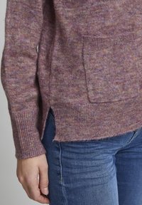 Lila kötött pulóver, textúrált felülettel, elülső zsebbel és oldalsó withslit-arral. Kék farmernadrággal párosítva.