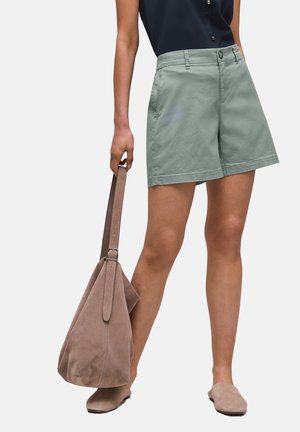 Kobieta w szortach w kolorze szałwi i czarnej bluzce, trzyma dużą torbę z zamszu w kolorze taupe i nosi pasujące taupe buty wsuwane.