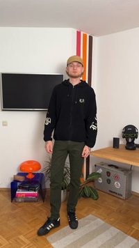 Felpa con cappuccio nera e zip frontale, pantaloni verdi e sneakers nere con strisce bianche; ambiente informale con decorazioni colorate.