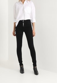 Černé skinny džíny s vysokým pasem, zavírání na pět knoflíků a hladkou texturou, v kombinaci s bílou košilí na knoflíky a černými botami.