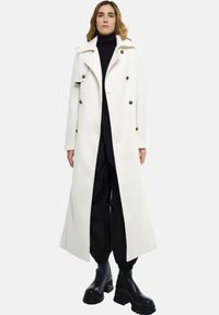 Long manteau blanc à double boutonnage, grands boutons et larges revers. Porté sur un col roulé noir et associé à des bottes noires.
