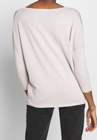 Pull en maille rose clair avec encolure bateau, manches 3/4 et texture côtelée. Présente une couture distincte dans le dos. Porté avec un pantalon foncé.