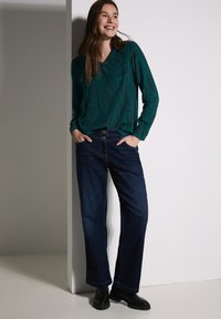 Blusa verde a motivi con maniche lunghe abbinata a jeans a gamba larga blu scuro e stivaletti neri alla caviglia, in piedi contro un muro bianco.