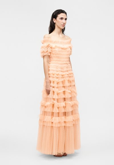 Naine kannab põlvini ulatuvat virsikuvärvi õlaosata kleiti kihiliste volangide ja läbipaistva kangaga, seisab ühevärvilise hele tausta ees.