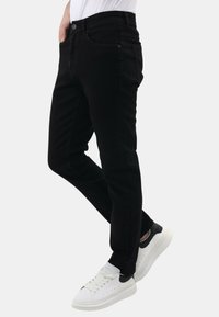 Persoon in slanke zwarte jeans en witte sneakers met zwarte hakken, staand met een iets gebogen been tegen een witte achtergrond.