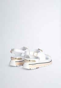 Sandali platform bianchi con accenti dorati, fibbia regolabile, suola testurizzata e dettagli borchiati sulle cinghie, posizionati su uno sfondo chiaro.