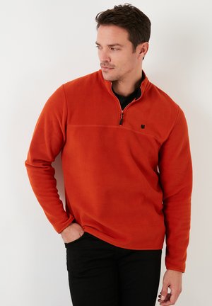 Pullover in pile arancione con zip a un quarto e logo nero sul petto. Maniche lunghe e una texture liscia, abbinato a pantaloni scuri.