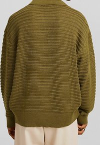 Personne portant un pull ample en maille vert olive avec des poignets et un ourlet côtelés, associé à un pantalon blanc cassé, vue de dos.