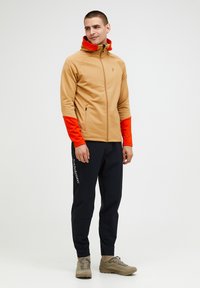 Giacca zip-up beige con maniche arancioni, tasche e cappuccio. Indossata con pantaloni neri con branding bianco. Il modello è in piedi in una postura rilassata.