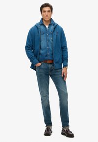 Sudadera azul superpuesta sobre una camisa de denim, combinada con jeans azules y zapatos de cuero marrón. Incluye capucha con cordón y bolsillos frontales.