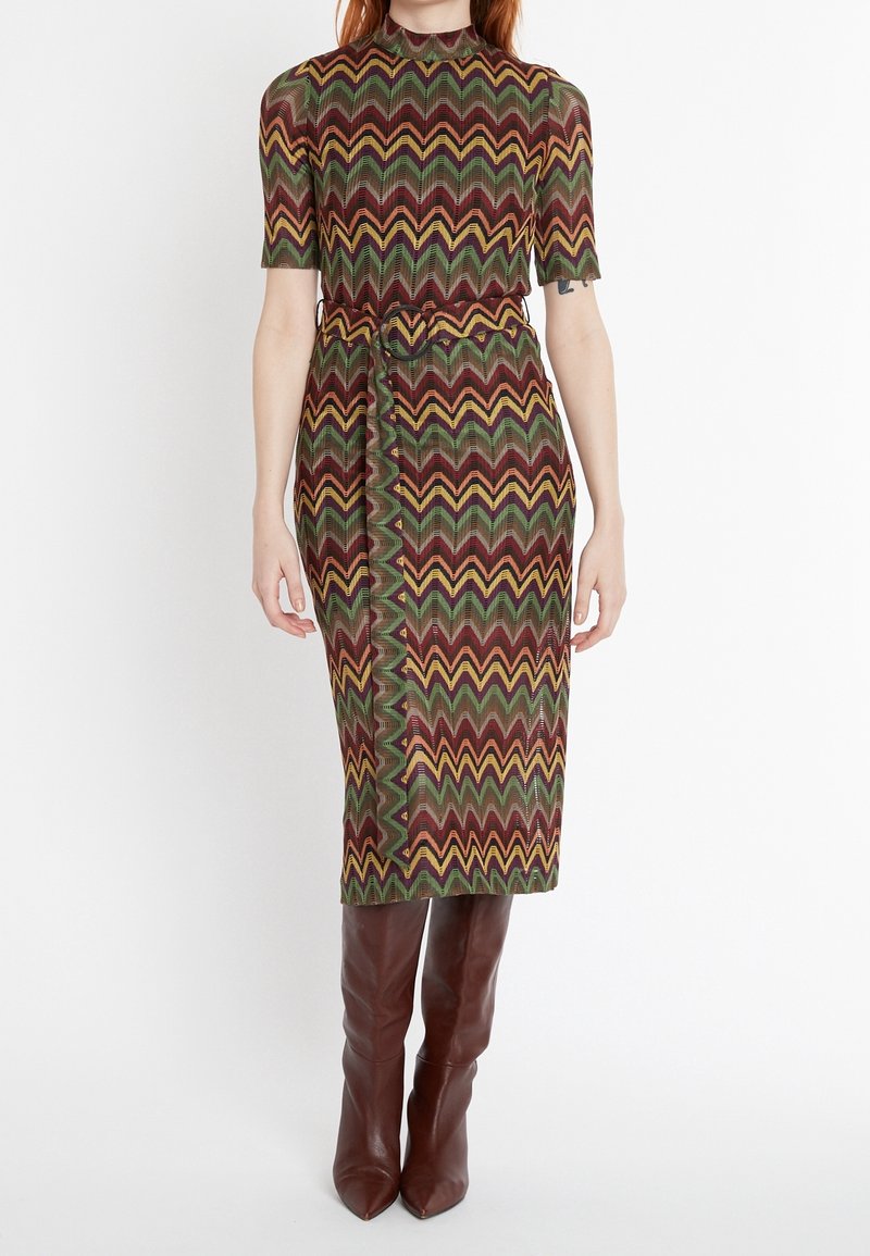 Sukienka do kolan z zygzakowatym wzorem w zielonym, brązowym i żółtym kolorze. Posiada krótkie rękawy, wysoką linię szyi oraz pasek w talii.