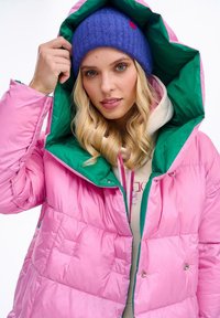 Nafouknutá růžová bunda se zelenou podšívkou kapuce, se zavíráním na patentky a prošívaným vzorem. V kombinaci s modrou pletenou čepicí.