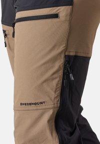 Beige och svarta utomhusbyxor med flera dragkedjefickor. Tillverkade av ett hållbart, texturerat tyg; har "SWEDEMOUNT"-logotypen nära fållen.