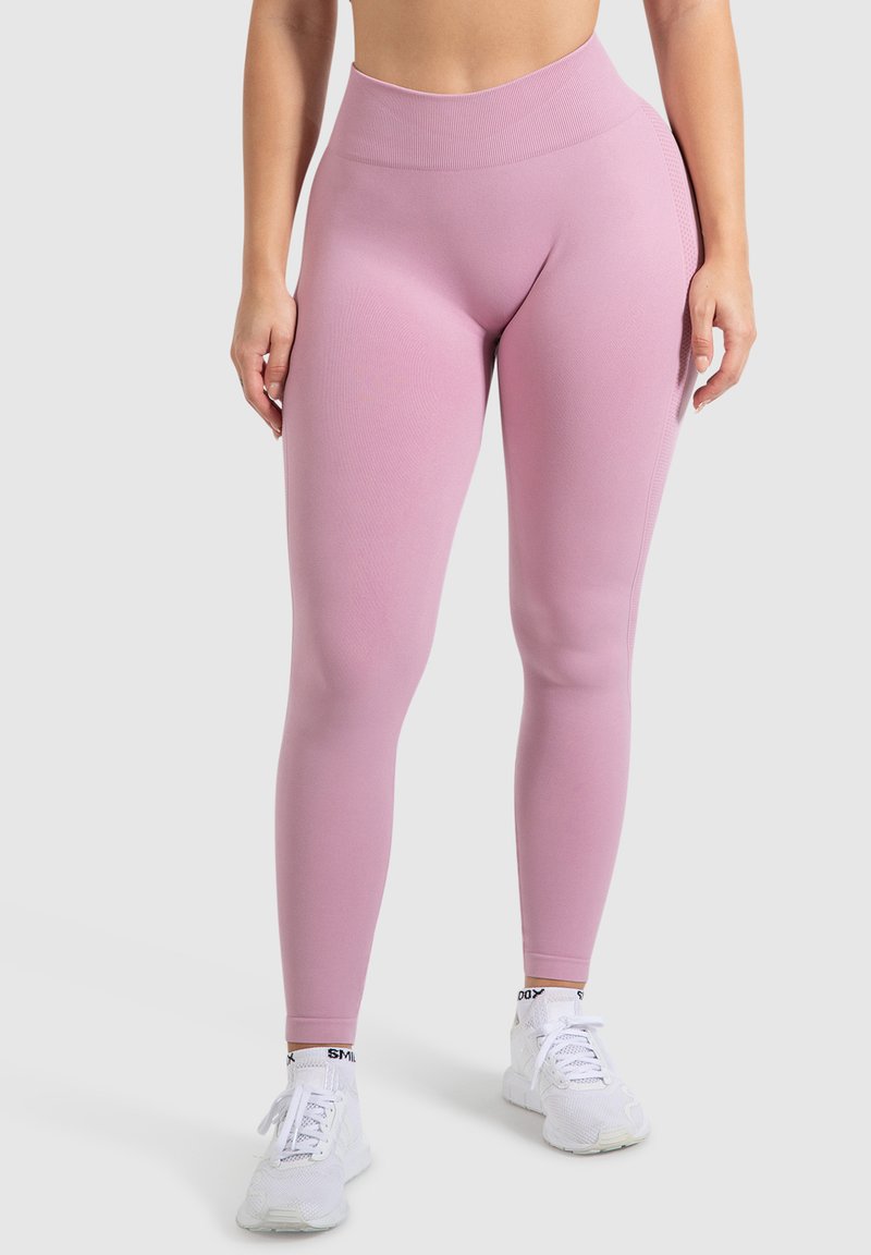 Leggings rosa a vita alta realizzati in tessuto elasticizzato, con una texture liscia e un design aderente, abbinati a scarpe da ginnastica bianche.