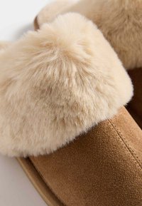 Pantofole in suede marrone con un collare in pelliccia sintetica color crema, caratterizzate da una texture morbida e un contrasto tra i materiali. Nessun hardware o dettaglio visibile.