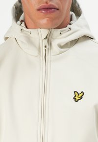 Primo piano di una persona che indossa una giacca con cappuccio di colore crema con un logo di un uccellino giallo sul petto e una chiusura lampo frontale.