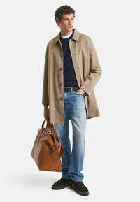 Beige bomullstrenchcoat med stora knappar, marinblå tröja, ljusblå jeans, brun läderväska och svarta skor. Enkelt, funktionellt design.