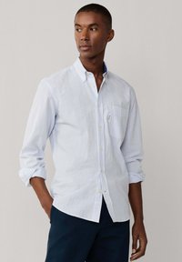 Homme portant une chemise à manches longues à rayures bleu clair et blanches, manches retroussées, debout avec une main dans la poche.