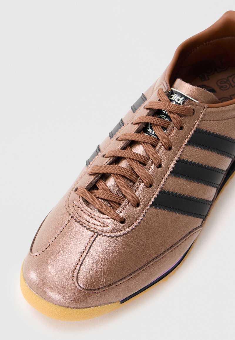 Sneaker in metallo bronzo con strisce nere, punta arrotondata e lacci marroni. Presenta una tomaia testurizzata e una suola in gomma naturale.
