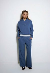 Niebieska bawełniana bluza z kapturem z motywem gwiazdy i kieszenią z przodu, w połączeniu z szerokimi niebieskimi spodniami. Oba elementy mają miękką fakturę i luźny krój.