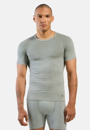 Mann mit kurzen Haaren, der ein enganliegendes hellgraues Sportshirt und Shorts trägt, steht frontal vor schlichtem Hintergrund.
