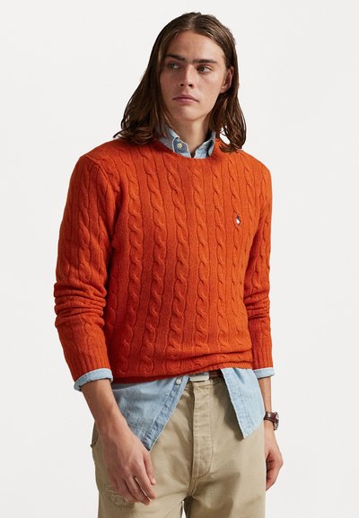 Pull en maille côtelée orange avec col rond, porté par-dessus une chemise en denim bleu clair. Détails de côtes remarquables et manches ajustées.