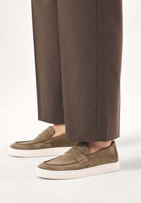 Mocassins en daim marron avec une finition texturée, comportant une lanière sur le dessus et une semelle en caoutchouc blanche. Associés à un pantalon marron sur mesure.