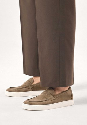 Mocasines de ante marrón con un acabado texturizado, que cuentan con una correa sobre la parte superior y una suela de goma blanca. Combinados con pantalones marrones a medida.