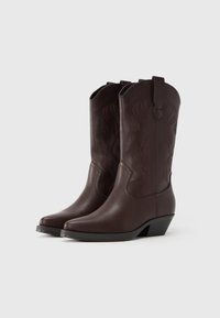 Barna műbőr cowboy csizma ívelt szegéllyel, részletes varrással és alacsony fekete sarokkal; sima textúra díszítő elemekkel.