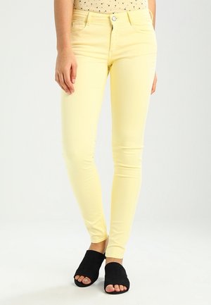 Kvinde iført lysegule slim-fit bukser og sorte åbne slip-on sko, der står mod en ensfarvet lys baggrund.