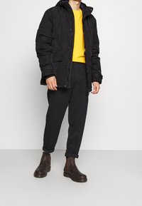 Černá prošívaná bunda s kapucí, žlutá mikina pod ní, černé zúžené kalhoty a hnědé kožené kotníkové boty. Neformální outfit.