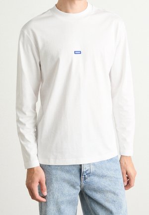 Mand iført almindelig hvid langærmet skjorte med lille blåt Hugo-logo på brystet og lyseblå denimjeans, stående mod hvid baggrund.