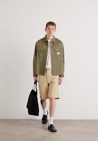 Grønn skjorte med knapper, beige shorts, svarte sko og hvite sokker; bærer en svart tote bag. Enkelt design, avslappet passform.