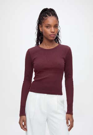Woman with long braided hair wearing a fitted maroon long-sleeve top and white high-waisted pants standing against a plain background.