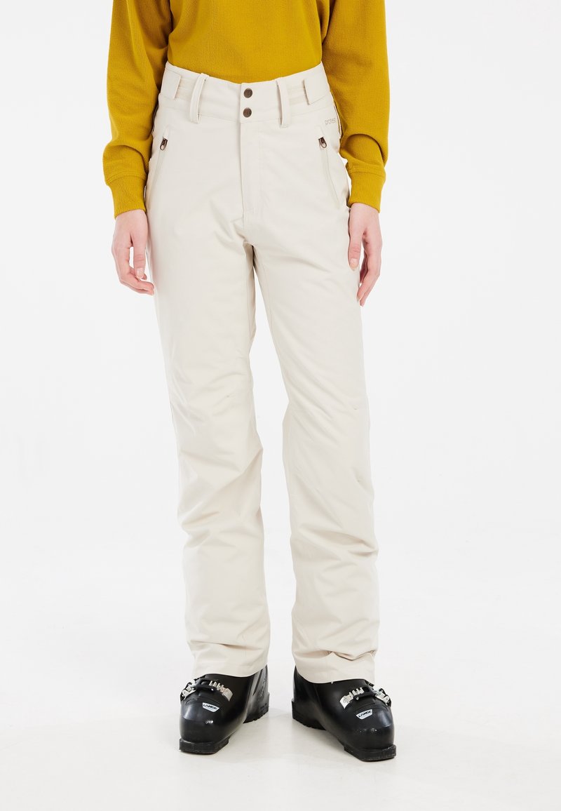 Personne portant un pantalon de ski beige, des bottes de ski noires et un haut à manches longues jaune moutarde, debout devant un fond blanc uni.