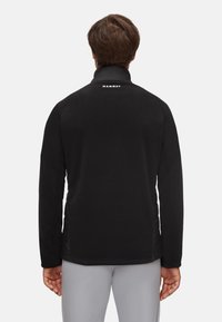 Hombre vestido con una chaqueta negra de Mammut con un cuello alto, visto desde atrás, de pie contra un fondo claro y liso.