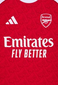 Rød stofftrøye med hevet mønster, hvit "Emirates Fly Better" logo og Adidas-logo i hvitt. Arsenal-klubbmerke er synlig.