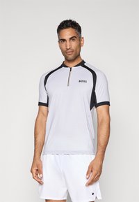 Mann in einem kurzärmeligen sportlichen Oberteil mit durchgehendem Reißverschluss in Schwarz und Weiß mit "BOSS"-Logo sowie weißen sportlichen Shorts, der vor einem einfarbigen Hintergrund steht.