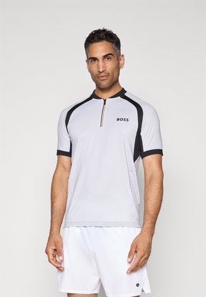 Mann in einem kurzärmeligen sportlichen Oberteil mit durchgehendem Reißverschluss in Schwarz und Weiß mit "BOSS"-Logo sowie weißen sportlichen Shorts, der vor einem einfarbigen Hintergrund steht.