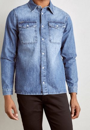 Mann trägt blaues Jeanshemd mit Brusttaschen und schwarzen Hosen vor einfarbigem Hintergrund.