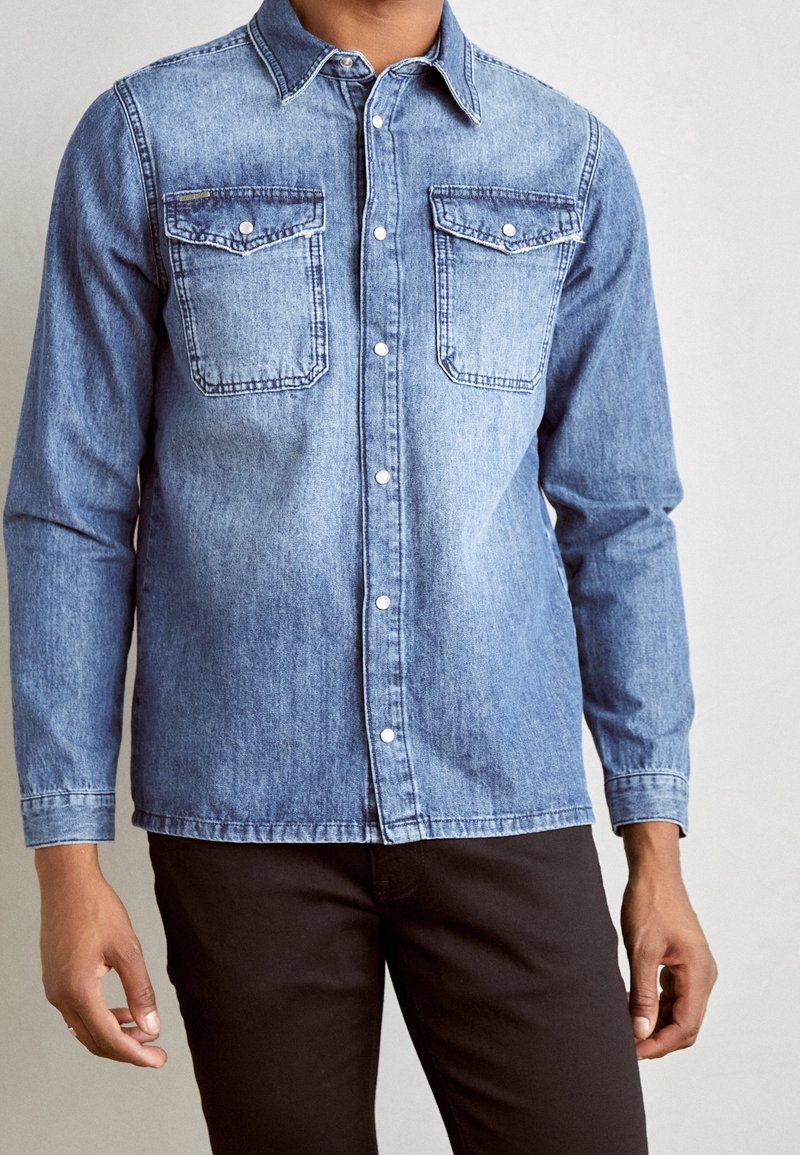 Homme portant une chemise en jean bleue à boutons avec poches poitrine et un pantalon noir, sur un fond uni.
