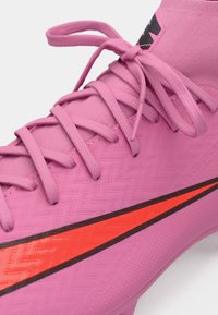 Roze voetbalschoenen met een gestructureerde oppervlakte, voorzien van roze veters, een opvallende rode en zwarte swoosh, en een kraagontwerp zoals een sok.