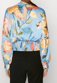 Kvinna i blommig blå blus med långa ärmar, smockad midja, knappstängning med nyckelhål bak och svarta byxor, sedd bakifrån.